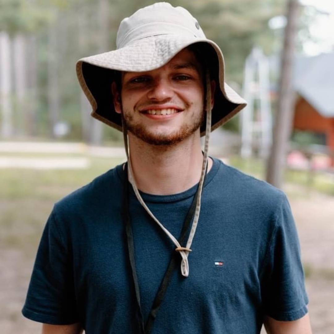 Image of a male smiling in a boat hat in the woods.